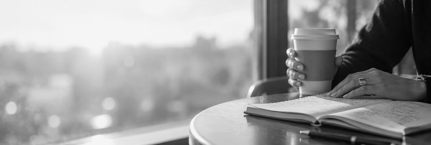 Hands holding a coffee cup over an open notebook at a sunlit window