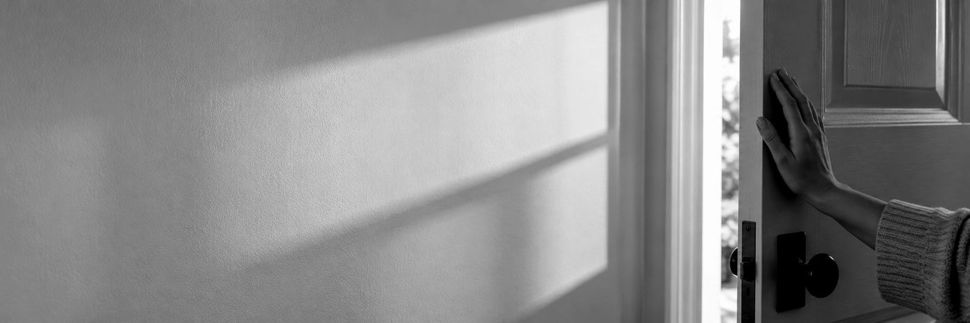 A woman's hand resting on the edge of a half-open front door in soft daylight